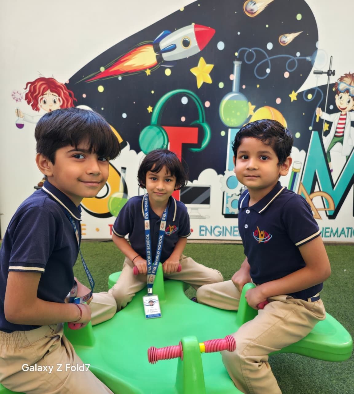 Three students on a green seesaw in front of a STEM mural